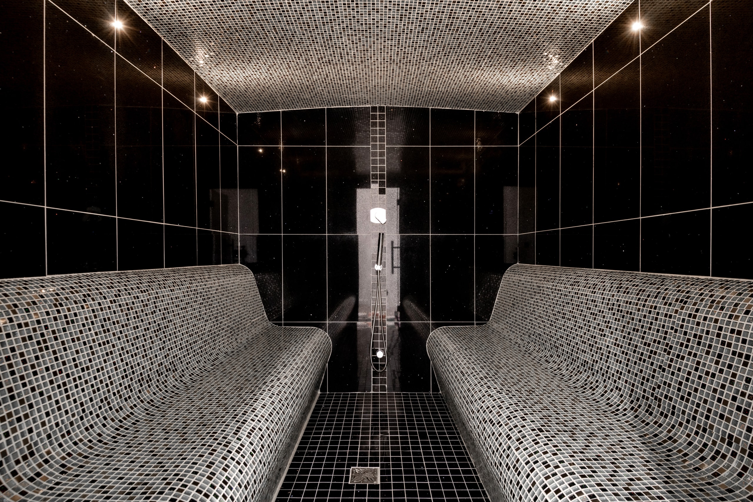Modern steam room with black tiled walls, gray mosaic seating benches, recessed lighting, and central shower fixture